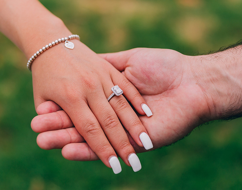 Image of a man and a woman holding hands outdoors. The woman has a diamond ring on her finger, and an elegant pendant bracelet